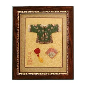 May include: Framed display featuring a green embroidered jacket with pink floral accents, a red tassel, and a pink fan. The items are set against a patterned background and framed in a dark wood frame.