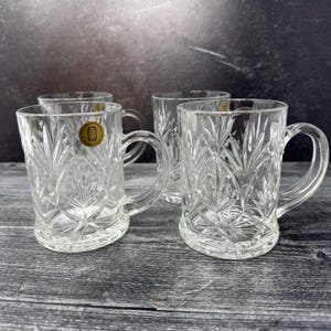 May include: Set of four clear glass mugs with a cut glass design. The mugs have a gold label with the text "Fine Crystal Made in USA".