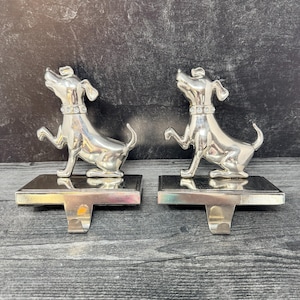 Puede incluir: Dos colgadores de calcetines plateados con forma de perro. Cada perro está en una pose juguetona, con un collar y una base rectangular con un gancho. Los perros miran hacia arriba. El fondo es gris oscuro.