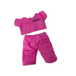 Könnte beinhalten: Ein hellrosa Puppen-Arztuniform-Set. Das Oberteil hat kurze Ärmel, eine Tasche und den Text "BABY LAND GENERAL HOSPITAL CLEVELAND GEORGIA". Die passende Hose ist in Capri-Länge.