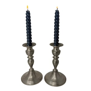 May include: Two antique silver candlesticks each hold a dark blue twisted candle with a lit yellow flame. The candlesticks have a classic design with a round base and a detailed stem.
