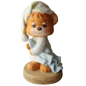 Puede incluir: Una figura de cerámica de Teddy Beddy Bear. El oso lleva un gorro de dormir y un pijama a rayas, sosteniendo una manta azul. El oso tiene mejillas rosadas y los ojos cerrados. La base de la figura es de color canela con la inscripción "Teddy Beddy Bear".