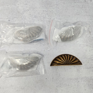 Könnte beinhalten: Vier antike Messing-Schrankknöpfe, drei davon in einzelnen durchsichtigen Plastiktüten und einer auf einer weißen Oberfläche. Zwei der Knöpfe sind halbrund und einer hat eine Fächerform.