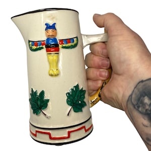 May include: Cream-colored ceramic pitcher with a black rim and handle. The pitcher features a colorful totem pole design and green maple leaf accents. The base has a red and black geometric pattern. The handle has a yellow and brown design.