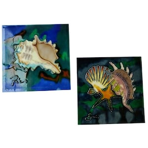 以下が含まれることがあります： 貝殻のデザインが施された2つの正方形のセラミックタイル。1つのタイルは、青と緑の背景に大きなクリーム色の巻貝が描かれています。もう1つのタイルには、ヒトデと貝殻が描かれています。