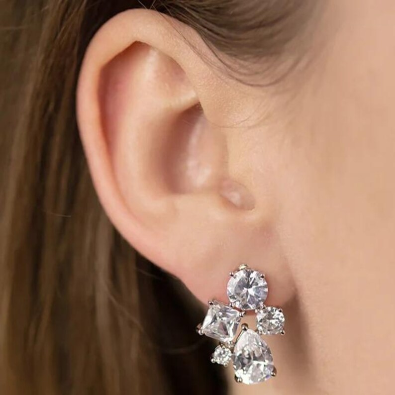 May include: A cluster earring featuring multiple clear, sparkling cubic zirconia stones. The earring has a round, square, and pear-shaped stones, set in a silver-toned metal. The earring is shown on an ear.