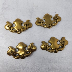 May include: Four gold-toned metal cabinet drawer pulls with a decorative, ornate design. Each pull has a curved, open handle.