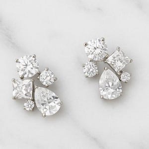 May include: A pair of silver-toned cluster earrings. Each earring features a collection of clear, sparkling cubic zirconia stones in various shapes, including round, pear, and square cuts. The earrings are set against a white marble background.