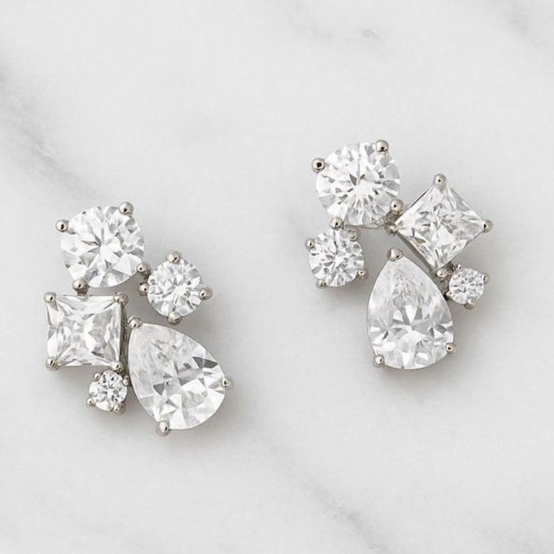 May include: A pair of silver-toned cluster earrings. Each earring features a collection of clear, sparkling cubic zirconia stones in various shapes, including round, pear, and square cuts. The earrings are set against a white marble background.