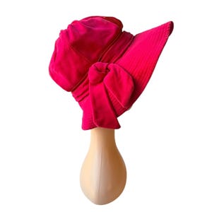 Puede incluir: Un sombrero de terciopelo fucsia vibrante con un gran lazo y un ala ancha. El sombrero se exhibe sobre una cabeza de maniquí sobre un fondo blanco. El diseño del sombrero incluye una corona estructurada y un lazo decorativo.