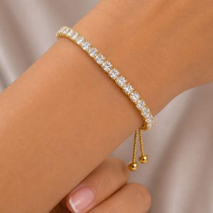 May include: A gold-toned bracelet with a row of square-cut, clear gemstones. The bracelet has a delicate design with a sliding clasp and two small gold spheres at the end of the adjustable chain. The bracelet is worn on a wrist.