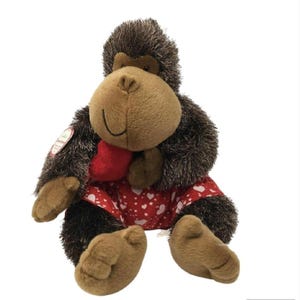 May include: A plush gorilla toy with brown fur, a tan face, and a red heart-shaped accent. The gorilla wears a red and white heart-patterned outfit. The toy is designed to sit upright.