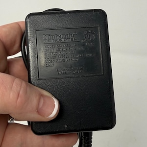 Puede incluir: Adaptador de CA negro de Nintendo con el texto "Nintendo AC Adapter" y "MODEL NO. NEB-00E" impreso en la parte delantera. El adaptador tiene un cable de alimentación negro.