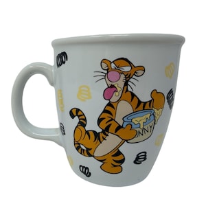 Puede incluir: Taza de cerámica blanca con una ilustración de dibujos animados de Tigger de Winnie the Pooh. Tigger, naranja con rayas negras, sostiene un tarro de miel. La taza tiene un asa curva y está decorada con garabatos amarillos y negros.