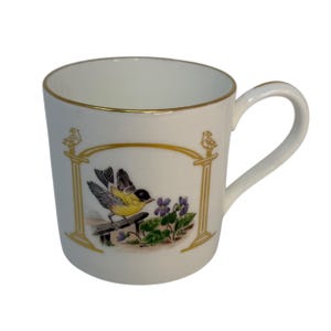 Può includere: Tazza in ceramica bianca con bordo e manico dorati. La tazza presenta un'illustrazione decorativa di un uccello giallo appollaiato su una staccionata di legno, con fiori viola e foglie verdi. Il design è incorniciato da accenti dorati.