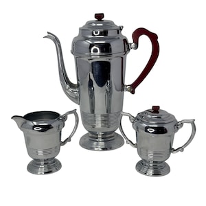 May include: A vintage chrome coffee set with a tall coffee pot, creamer, and sugar bowl. The coffee pot has a red handle and lid finial. The set has a reflective surface and a ribbed design on the base.