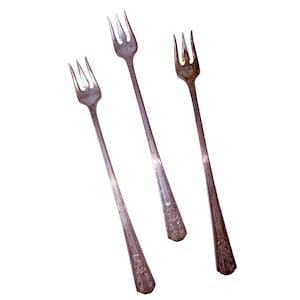 May include: Three antique silver forks with long, slender handles and three tines. The forks have decorative detailing at the base of the handle. The forks are arranged diagonally against a white background.