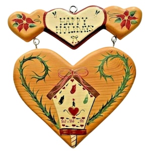 May include: Wooden heart-shaped Christmas decoration with a birdhouse design. The birdhouse is painted with festive symbols and the words "Ho Ho Ho". The top heart reads "Happy Holidays". The decoration is accented with red and green details.