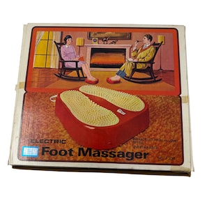 Pode incluir: Caixa de massajador de pés elétrico vermelho vintage com uma imagem de um casal relaxando. O massajador tem almofadas texturizadas amarelas. O texto na caixa diz "Electric Foot Massager". A caixa é branca e tem um design retrô.