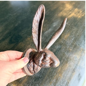 May include: A cast iron wall hook shaped like a rabbit head with long, upright ears. The object features a textured, dark brown, rustic finish with visible metal casting details and a circular base for mounting against a flat surface.