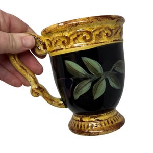 May include: A decorative ceramic mug with a black body, adorned with a green leaf design. The rim and handle feature a yellow and brown ornate pattern. The mug is held by a hand against a white background.