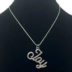 Może przedstawiać: Srebrny naszyjnik z wisiorkiem z napisem "Joy" w ozdobnej czcionce. Wisiorek jest inkrustowany małymi, błyszczącymi kryształkami. Naszyjnik wisi na czarnym tle, podkreślając jego design.