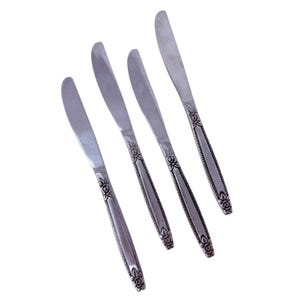 May include: Four silver butter knives with rounded blades and decorative floral patterns on the handles. The knives are arranged diagonally against a white background, showcasing their polished surfaces and intricate design.