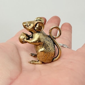 May include: A small, detailed brass mouse figurine holding a nut. The mouse has a curled tail and detailed features, with a slightly aged appearance. The figurine is a decorative item.