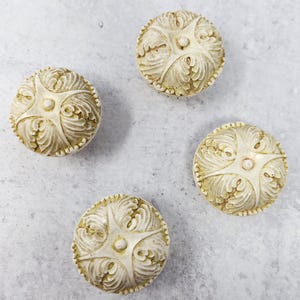 Könnte beinhalten: Vier cremefarbene Keramik-Schrankknöpfe mit einem verzierten, erhabenen Blumenmuster. Die Knöpfe haben einen leicht vergilbten Farbton.