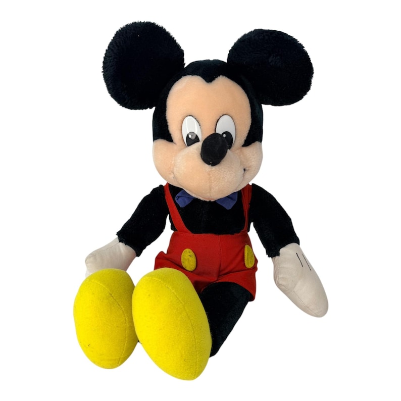 Peut inclure: Une peluche Mickey Mouse avec des oreilles noires, un corps noir et un visage beige. Mickey porte une salopette rouge avec des boutons jaunes, des gants blancs et des chaussures jaunes. Un n&oelig;ud papillon bleu compl&egrave;te le look classique.
