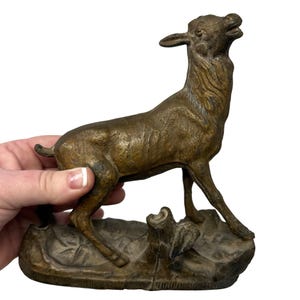 May include: A bronze cast figurine of a deer standing on a base with a tree stump. The deer is looking up and has its mouth open.