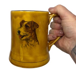 Puede incluir: Taza de cerámica de color amarillo mostaza con un retrato detallado de un perro con pelaje marrón y canela. La taza tiene un asa curvada y un borde marrón oscuro. El perro está sonriendo con la lengua fuera.