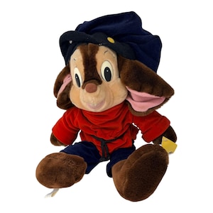 Peut inclure: Peluche d'un personnage de dessin animé, avec un visage brun et beige, de grands yeux et un petit nez. Le personnage porte un chapeau bleu marine, une chemise rouge et un pantalon bleu. Le jouet a de grands pieds et oreilles bruns et poilus. Une étiquette jaune est attachée.