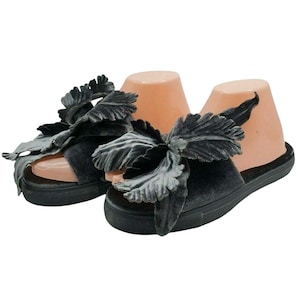 May include: Pair of dark gray velvet slide sandals. Each sandal features a large, decorative leaf-shaped embellishment. The sandals have a thick, black sole and are displayed on a mannequin foot form.