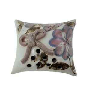 May include: A ceramic, pillow-shaped air freshener with a white base. It features a raised pink bow and floral designs in pink and blue. The design includes several small holes.