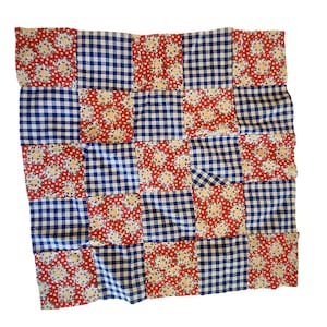 Op de afbeelding: Een patchworkquilt met een herhalend patroon van rode bloemen vierkanten en blauw-witte ruitjes vierkanten. De quilt is gemaakt van stof en heeft een handgemaakt uiterlijk. De quilt is vierkant van vorm.