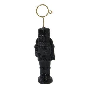 Puede incluir: Figura de cascanueces negra con un lazo de alambre dorado para sujetar notas o fotos. La figura está detallada con un uniforme y un sombrero. La base es redonda. El fondo es blanco.