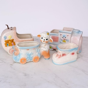 Könnte beinhalten: Eine Sammlung von Vintage-Keramik-Baby-Themen-Pflanzgefäßen in Pastellfarben. Enthält einen Kinderwagen, eine Lokomotive, einen Stiefel und eine Lammfigur. Auf der Lokomotive steht "By Land Express". Die Pflanzgefäße sind mit Blumen- und Tiermotiven verziert.