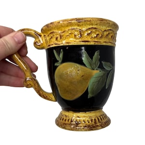 May include: A ceramic mug with a black background and a yellow pear design. The mug has a decorative yellow rim and base, and a curved handle. The pear is painted in shades of yellow and green, with green leaves. The mug is held by a hand.