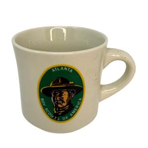 Könnte beinhalten: Weiße Keramiktasse mit einem grün-gelben Logo der Boy Scouts of America, das einen Pfadfinder mit Hut und dem Text "Atlanta" zeigt.