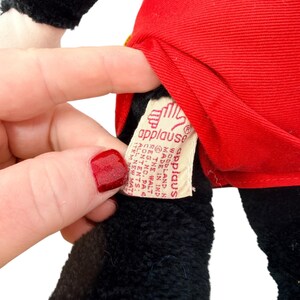 Peut inclure: Gros plan sur un jouet en peluche avec une veste rouge et de la fourrure noire. Une &eacute;tiquette avec le logo "applause" est visible. L'&eacute;tiquette comprend &eacute;galement du texte sur les mat&eacute;riaux et l'origine du jouet.