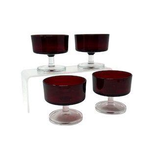 Peut inclure: Quatre verres à pied vintage de couleur bordeaux. Les verres ont un bol large et peu profond et une tige transparente et courte. Ils sont disposés sur une étagère transparente et incurvée sur fond blanc. Les verres sont probablement destinés à servir des desserts ou des cocktails.