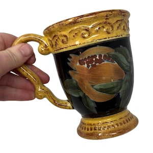 May include: A ceramic mug with a black background and a painted orange flower design. The rim and base of the mug are yellow with brown accents. The handle is also yellow. The mug is held at an angle.