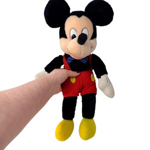 Peut inclure: Poup&eacute;e en peluche Mickey Mouse avec des oreilles, des bras et des jambes noirs, un visage cr&egrave;me et une tenue de short rouge. La poup&eacute;e a un n&oelig;ud papillon bleu, des gants blancs et des chaussures jaunes. C'est un personnage Disney classique.