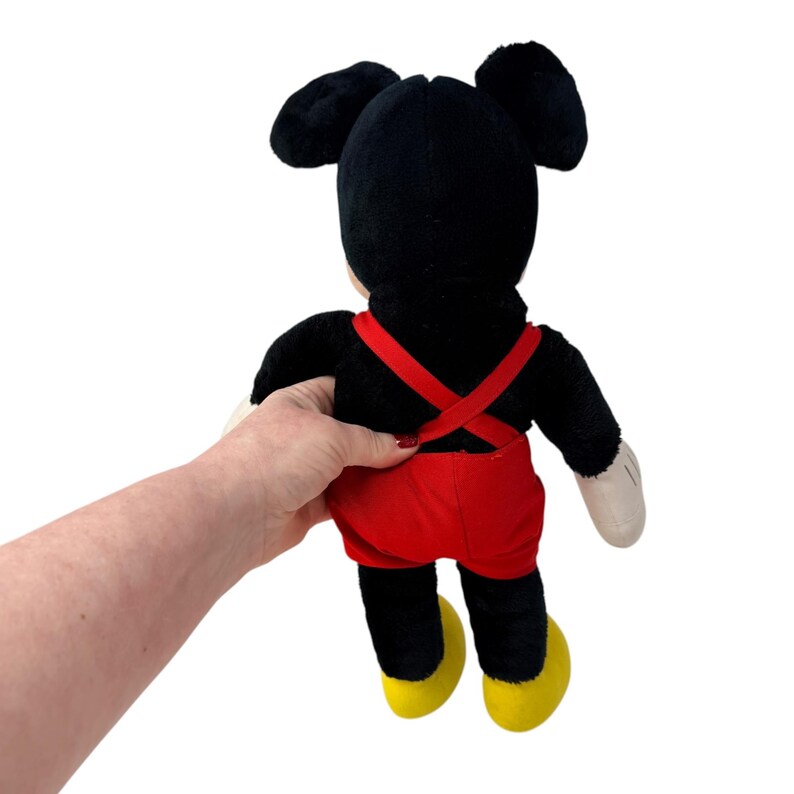 Peut inclure: Une poup&eacute;e en peluche de Mickey Mouse, vue de dos, tenue par une main. La poup&eacute;e pr&eacute;sente une fourrure noire, un short rouge avec des bretelles, des chaussures jaunes et des gants blancs. Les oreilles embl&eacute;matiques du personnage sont bien visibles.