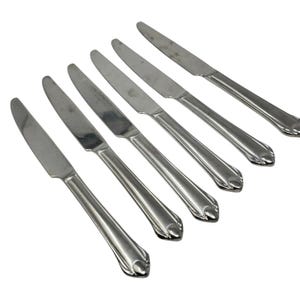 May include: Six stainless steel dinner knives arranged diagonally. Each knife has a smooth, shiny blade and a handle with a decorative, curved design at the end. The knives are set against a white background, highlighting their metallic sheen.