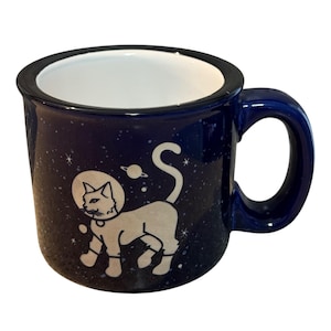 Puede incluir: Una taza de cerámica azul oscuro con interior blanco y asa. La taza presenta una ilustración blanca de un gato con traje espacial, con un diseño de planeta y estrellas. El borde y el asa son de color azul oscuro.