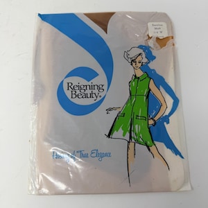Puede incluir: Un paquete de medias Reigning Beauty. El embalaje es transparente con un diseño azul y blanco y las palabras "Reigning Beauty" en un círculo azul. Una ilustración de una mujer con un vestido verde está en el embalaje. También son visibles las palabras "Hosiery of True Elegance".