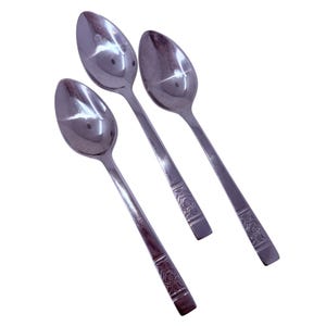 Puede incluir: Tres cucharas de acero inoxidable con acabado pulido. Cada cuchara tiene un cuenco redondeado y un mango largo con un patrón floral decorativo cerca del extremo. Las cucharas están dispuestas en diagonal sobre un fondo blanco.