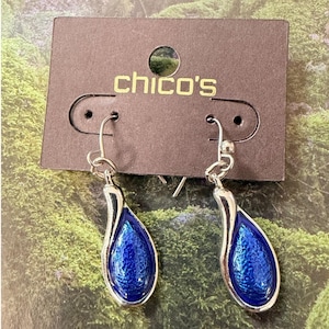 Puede incluir: Un par de pendientes colgantes de tono plateado con un colgante en forma de lágrima y una incrustación azul metálica texturizada. Los pendientes están sujetos a una tarjeta de cartón marrón con la marca chico's impresa en letras doradas.